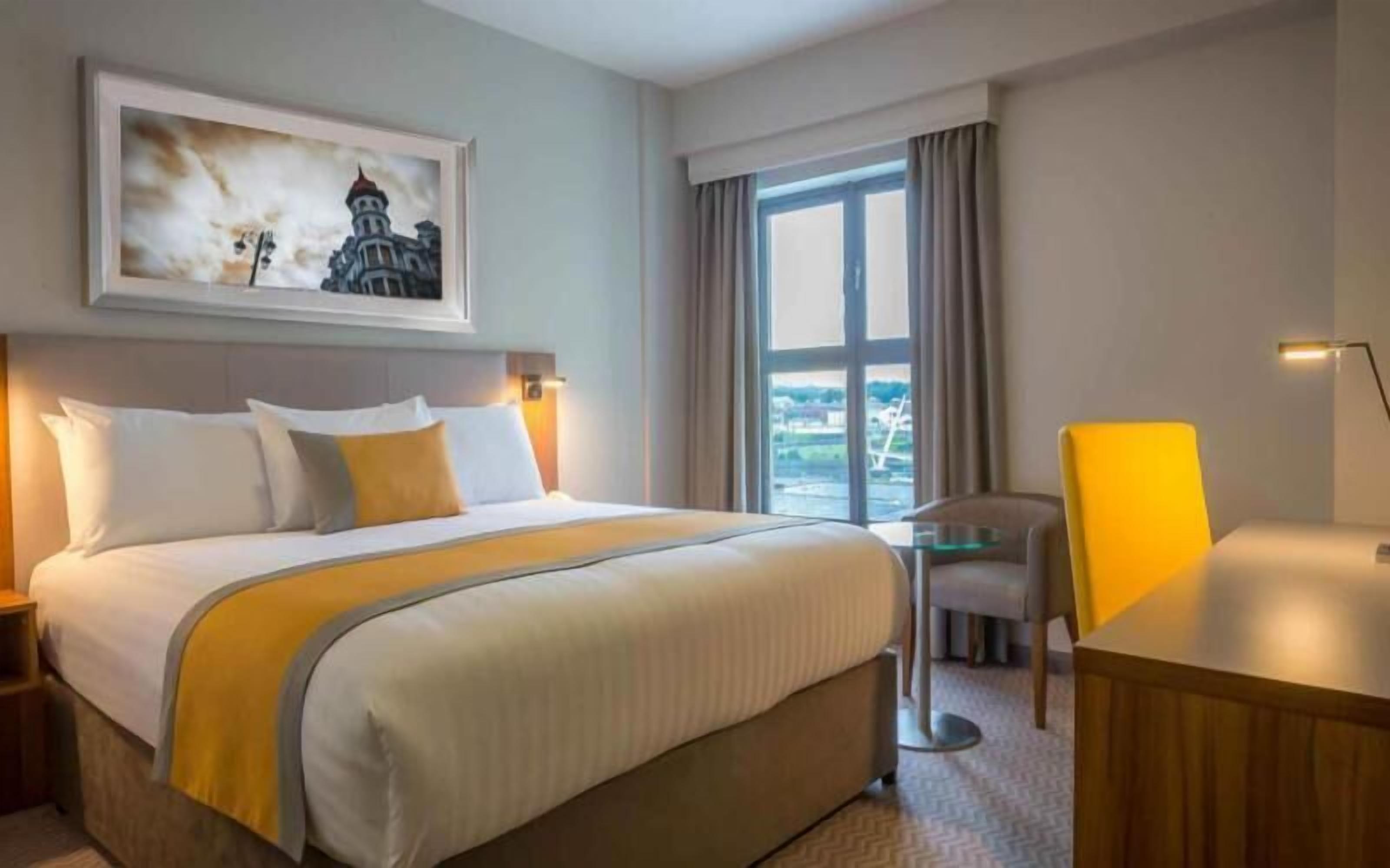 Habitación de hotel con cama, escritorio, silla, ventana con vistas y un cuadro en la pared.