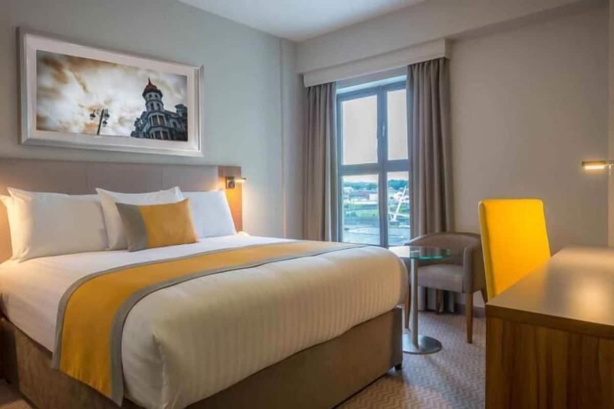 A hotel room with a bed, a desk, a chair, a window with a view, and a painting on the wall.