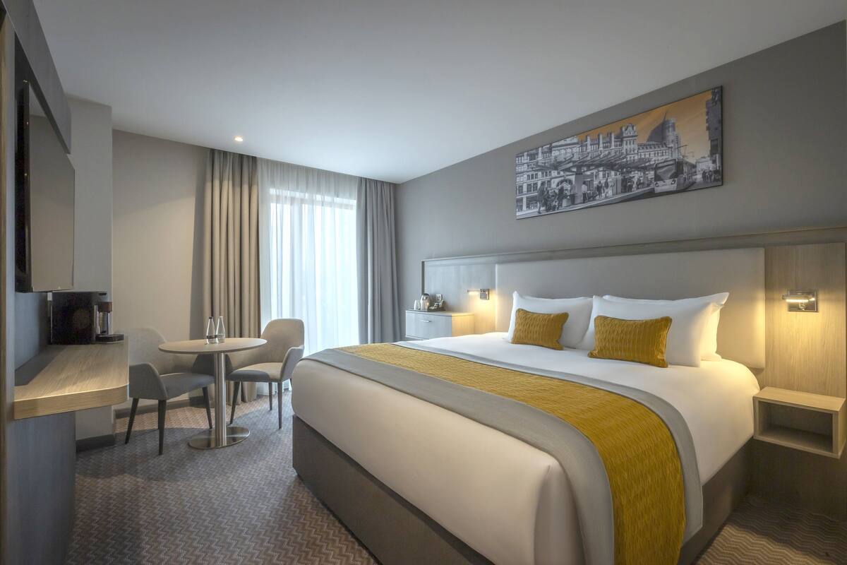 A modern hotel room with a large bed, a desk with two chairs, a small table, and a painting on the wall.