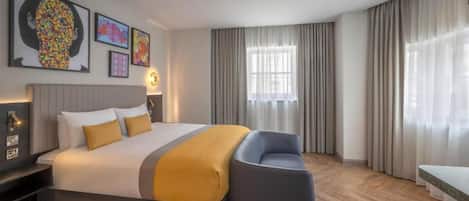 Una habitación de hotel moderna con una cama grande, una mesita de noche, una silla, una ventana con cortinas y una obra de arte en la pared.