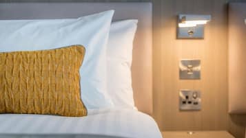 Una cama bien hecha con almohadas blancas y doradas, una mesita de noche con lámpara, e interruptor de luz en la pared.