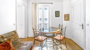 Un comedor con una mesa redonda de vidrio y sillas metálicas, un sofá estampado y vista a un edificio a través de una ventana con cortinas.