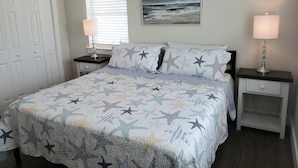 A bedroom with a bed, two bedside tables, a nightstand, a door, a window with blinds, and a painting on the wall.
