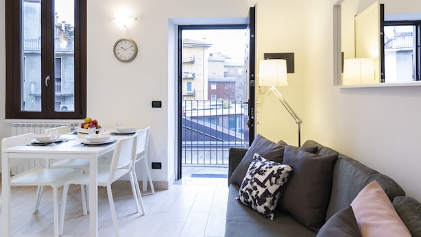 Una sala moderna con un sofá gris, una mesa de comedor blanca y sillas. Un reloj está en la pared, y una puerta da a un balcón exterior.