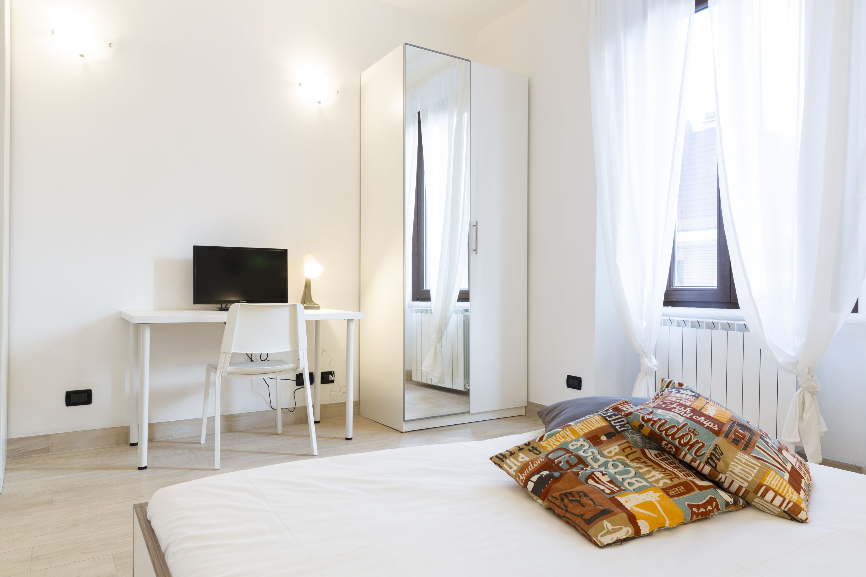 A bedroom with a bed, a desk with a chair, a television, a wardrobe, and a window with curtains.