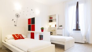 Un dormitorio moderno con dos camas, un armario blanco, una mesita de noche con lámpara y una ventana con cortinas de gasa.