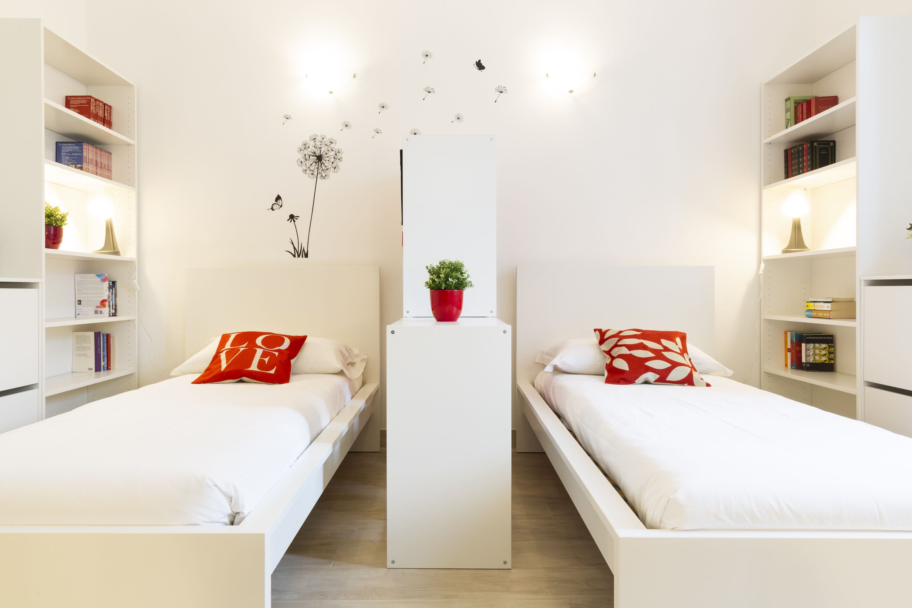 A modern hotel room with two single beds, white bedding, and red pillows. There are white shelves on either side of the room with books and decorative items. A central white cabinet is placed between the beds.