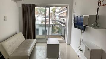 Una sala de estar moderna con sofá, una mesa de centro, un televisor de pantalla plana montado en la pared y vistas a una zona residencial a través de amplios ventanales.