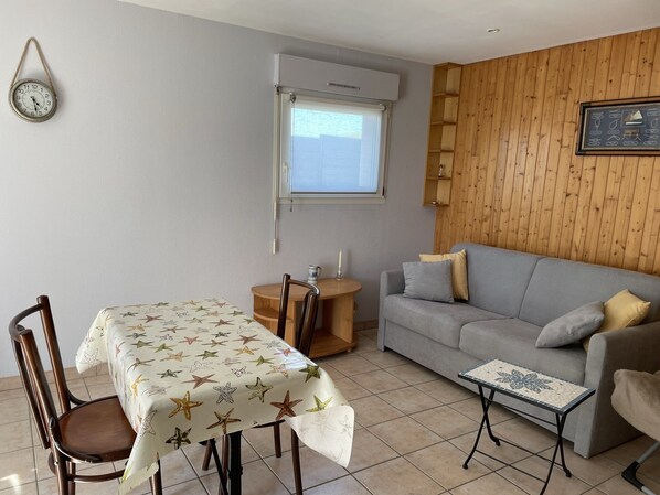 Un salon avec un canapé gris, une petite table recouverte d’une nappe à motifs, une chaise en bois, une horloge au mur et une fenêtre avec des stores.
