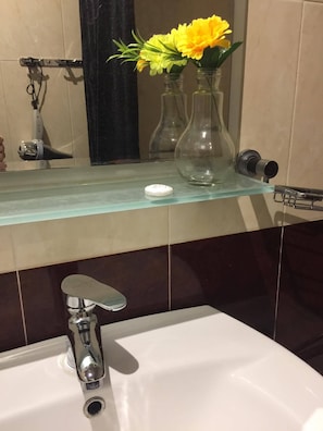 A bathroom with a glass shelf holding two clear vases with yellow flowers, a white sink with a chrome faucet, and a towel rack with a black towel. - Suit apartment Erekle Tatishvili street (Tbilisi)