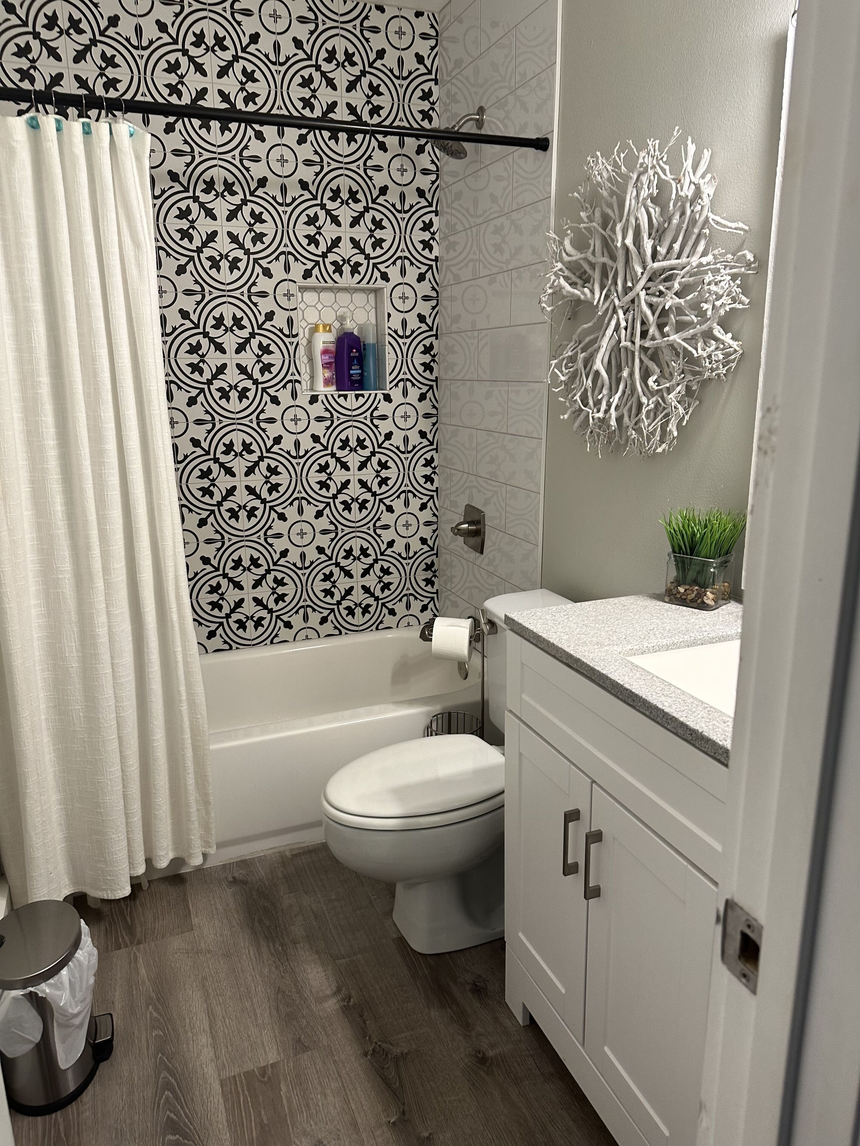 A bathroom with a patterned wall, a toilet, a bathtub, a shower curtain, a sink with a decorative piece, and a trash bin.