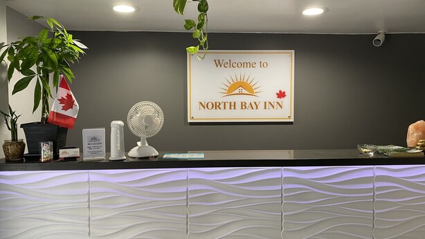 A reception desk at North Bay Inn with a welcome sign, a fan, a potted plant, and a Canadian flag. - North Bay Inn (North Bay)