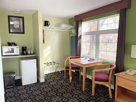 Habitación de hotel compacta con kitchenette, un comedor con dos sillas, un refrigerador pequeño, microondas, cafetera, teléfono y ventana con vista a árboles.