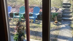Blick durch ein Fenster auf einen Patio mit zwei blauen Adirondack-Stühlen, einem Blumentopf und einer Steintreppe, die zu einer Terrasse führt.