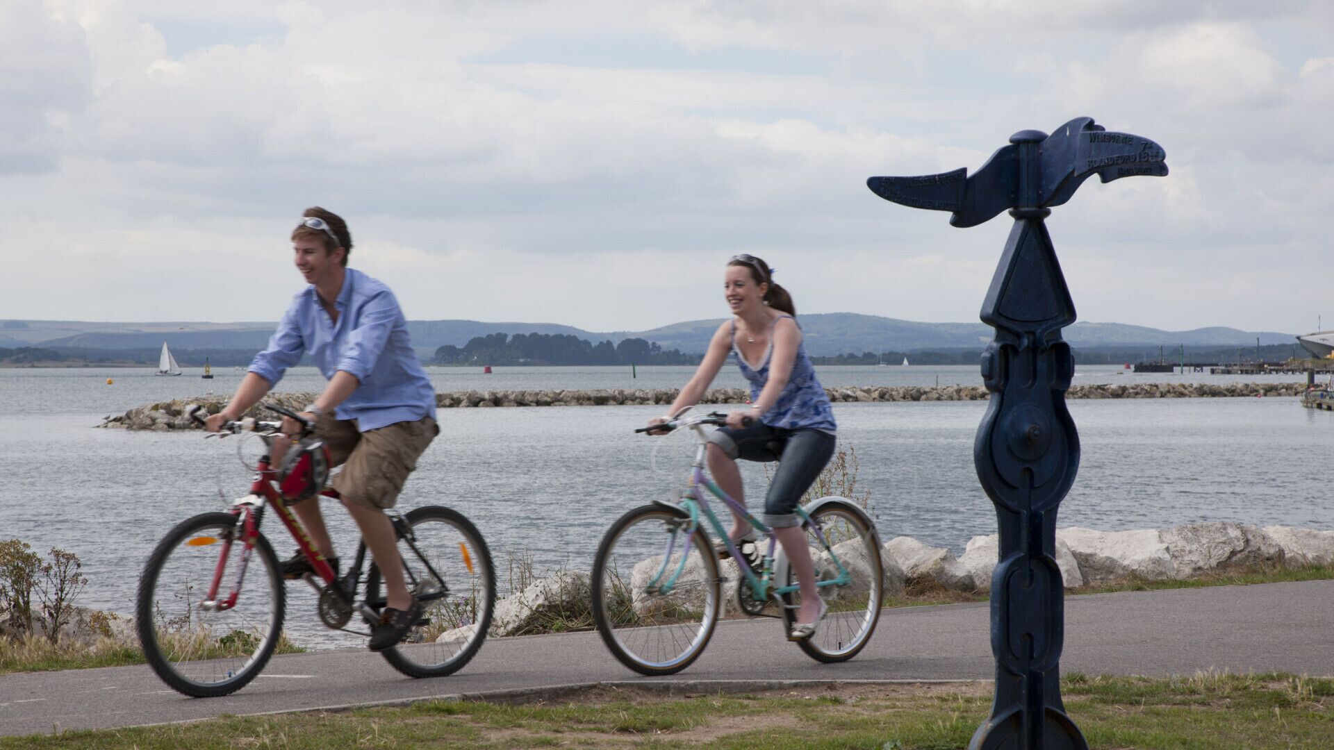 Dos personas montando en bicicleta por un camino pavimentado junto a una masa de agua con costa rocosa y colinas al fondo.