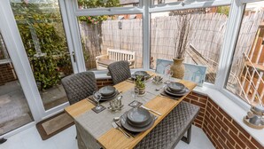 Un coin repas avec une table dressée pour quatre personnes, entouré de chaises confortables, d’une table en bois et offrant une vue sur un jardin grâce à de grandes fenêtres.