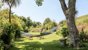 Un hamac tendu entre deux arbres, dans un jardin luxuriant, avec un banc en pierre et une maison en arrière-plan.