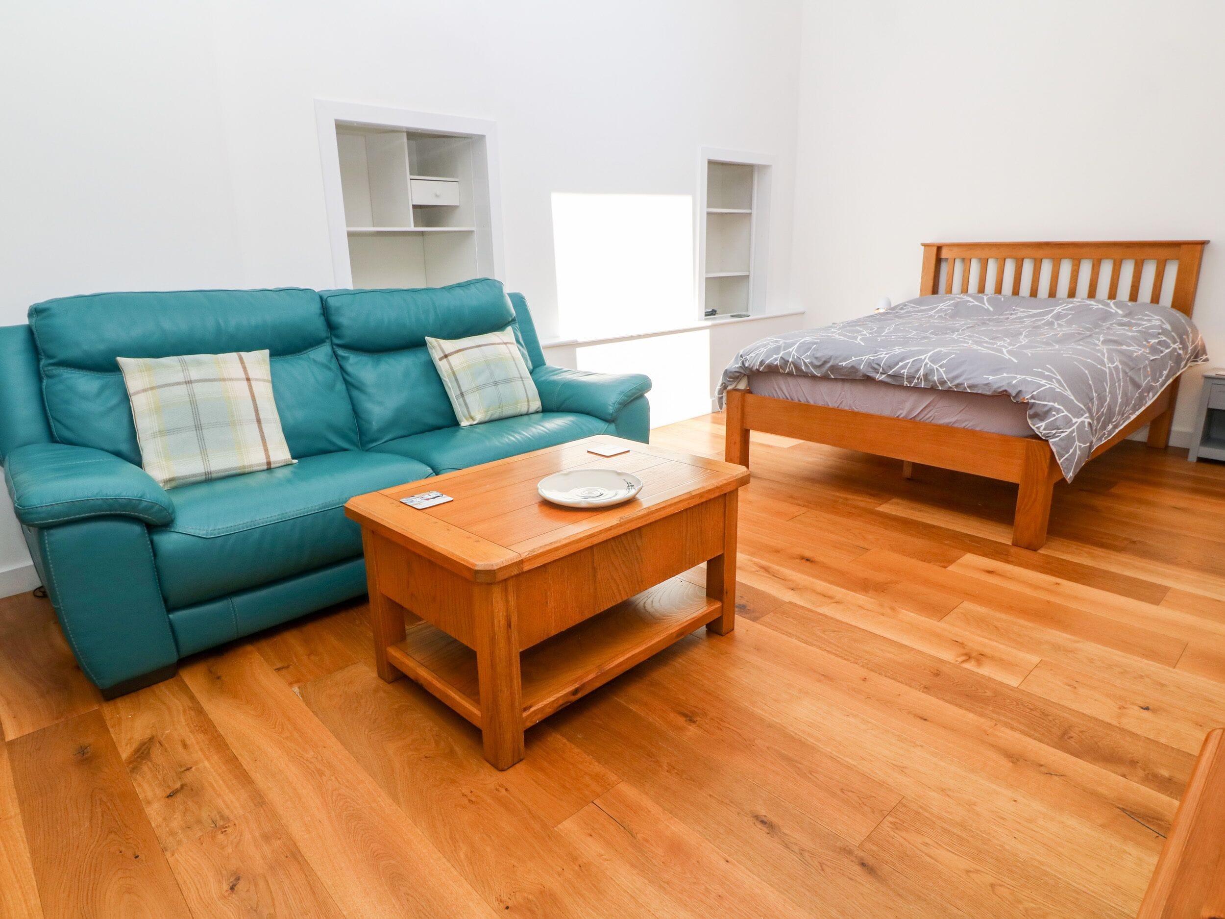 Una habitación con un sofá color turquesa, una mesa de centro de madera, una cama de madera con cubrecama gris y un armario blanco.