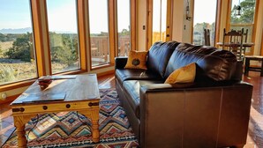 Un salón espacioso con un sofá de cuero, una mesa de centro de madera y una alfombra colorida. Grandes ventanales ofrecen vistas al exterior.