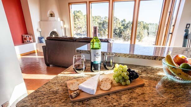 A kitchen counter with a bottle of wine, glasses, cheese, grapes, and a bowl of fruit. - Dark Sky Retreat Near Santa Fe (Santa Fe)