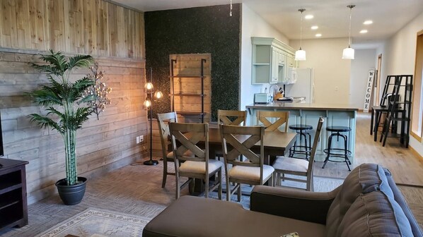 Una sala de estar moderna con una pared de acento de madera, un comedor con mesa y sillas de madera, una cocina con armarios blancos y una zona de bar con taburetes.