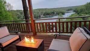 Une terrasse dotée de deux fauteuils confortables et d’une table carrée avec un foyer intégré, offrant une vue sur une rivière et une zone boisée.