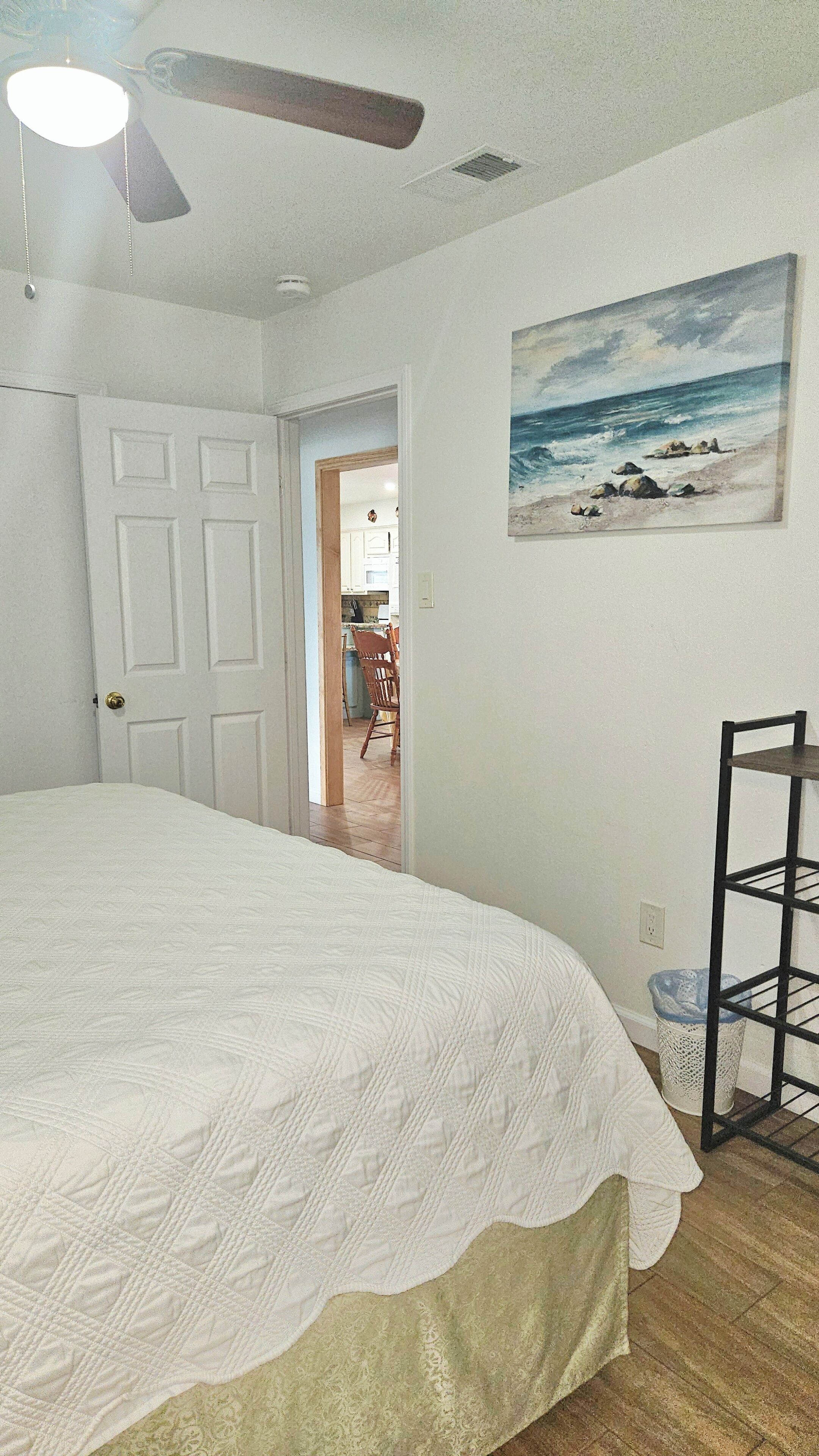 Un dormitorio con cama, ventilador de techo, un cuadro de playa y un pasillo que conduce a la cocina.