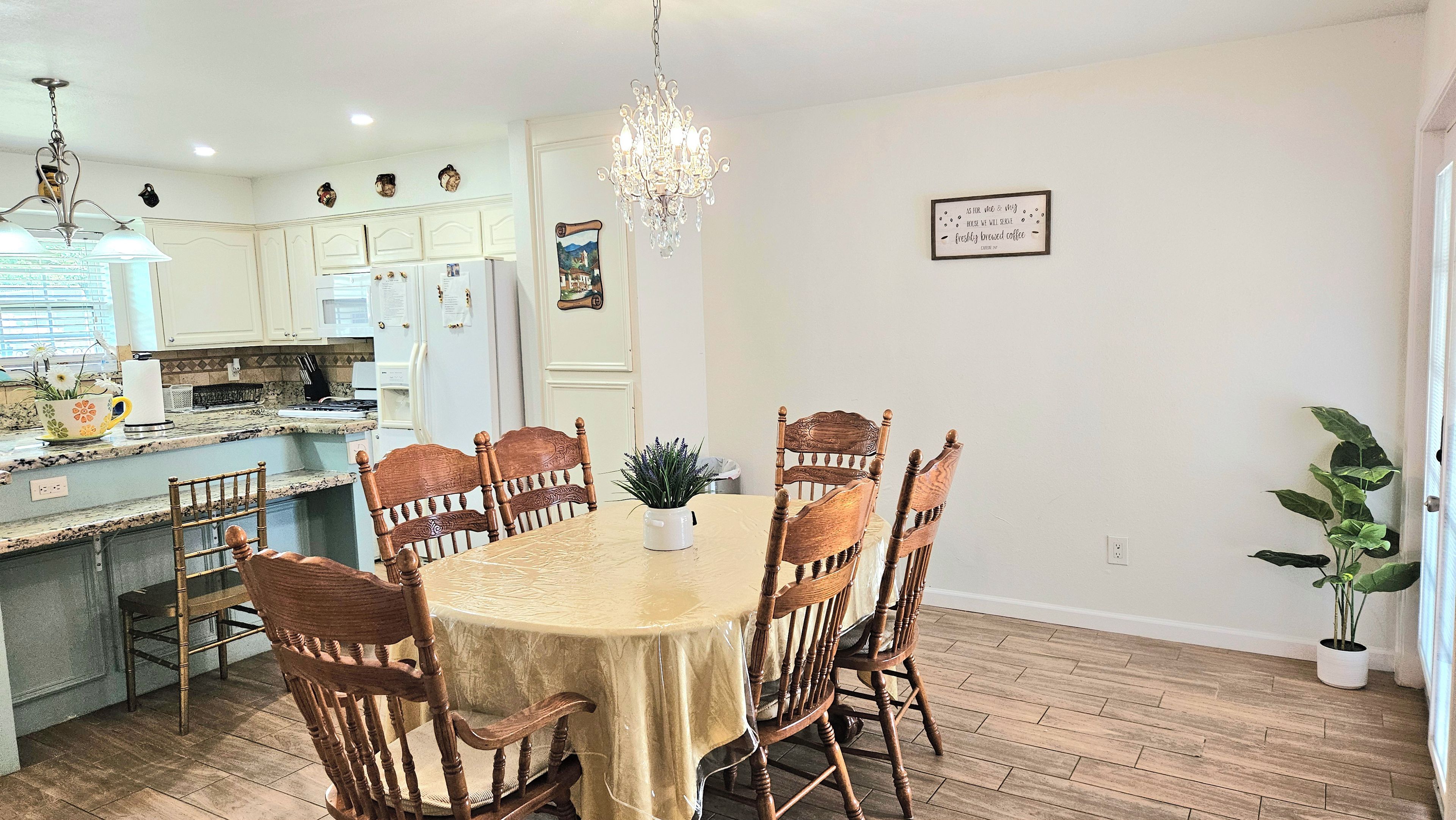 A dining area with a round table, six wooden chairs, a chandelier, and a kitchen in the background.