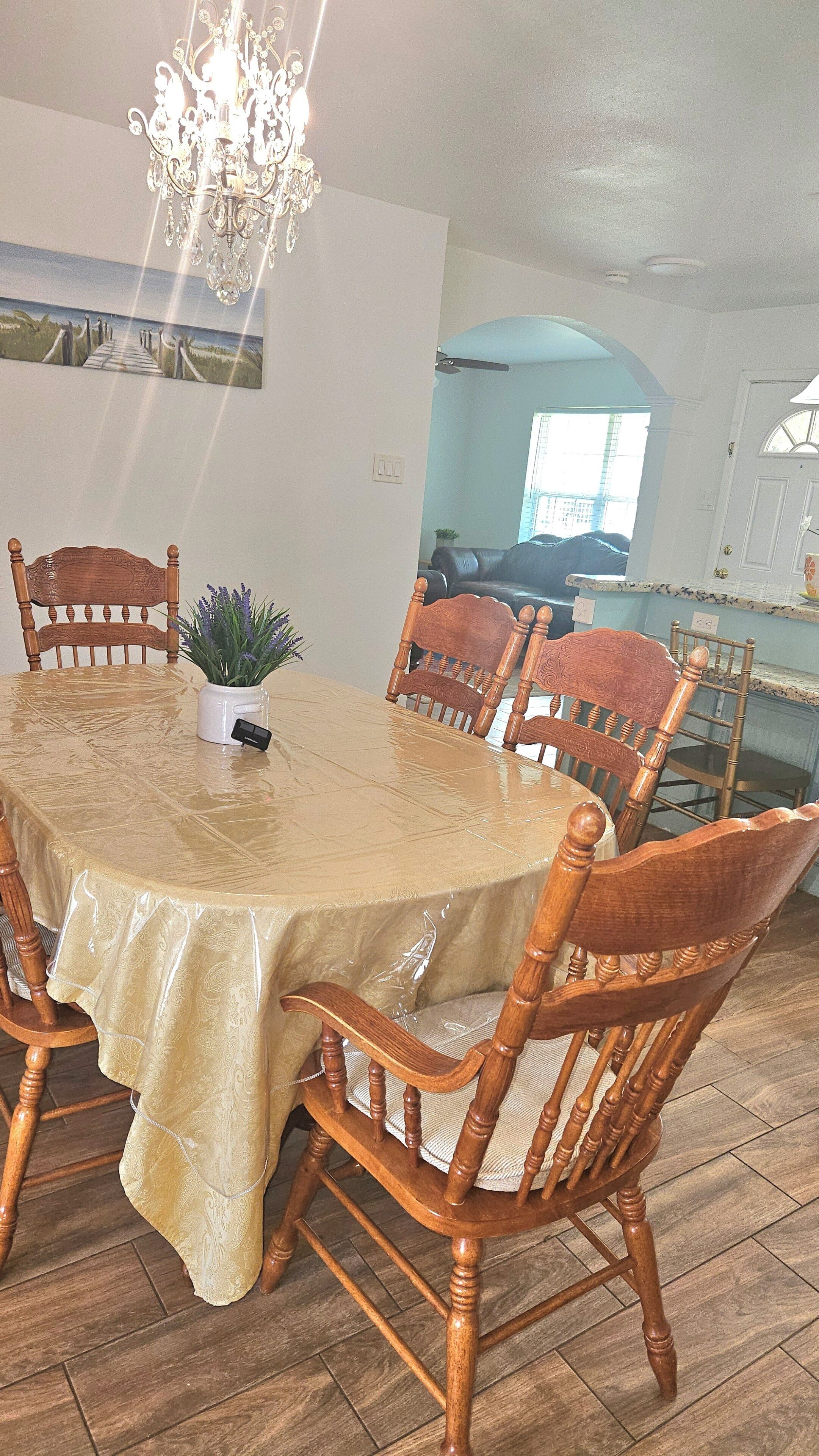 Un comedor con una mesa cubierta con mantel color beige, rodeada de sillas de madera. Un candelabro cuelga del techo, y se aprecia un cuadro con una escena de playa en la pared.