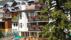 Ein mehrstöckiges Hotel mit Balkonen, die mit bunten Blumenkästen geschmückt sind, eine Steintreppe und Blick auf eine Berglandschaft.