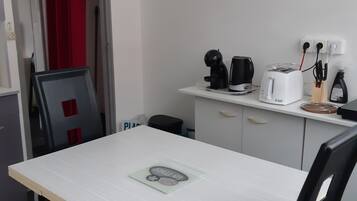 Une petite pièce propre, avec une table blanche, des chaises noires, un rideau rouge, une bouilloire noire, un grille-pain blanc et une petite poubelle.