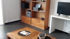 Ein Wohnzimmer mit Holztisch, einer Couch, einem Fernseher auf einem Schrank und einem Bücherregal mit verschiedenen Gegenständen.