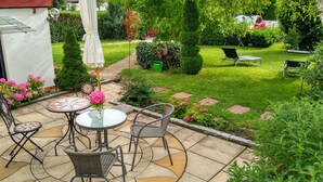 A patio area with a glass table and chairs, surrounded by a well-maintained garden with hedges and flowering plants. - Small 3-room apartment with large terrace, 150m to the lake (Uhldingen-Mühlhofen)