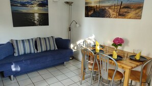 A living room with a blue sofa, a dining table with chairs, and two wall-mounted paintings. - Small 3-room apartment with large terrace, 150m to the lake (Uhldingen-Mühlhofen)