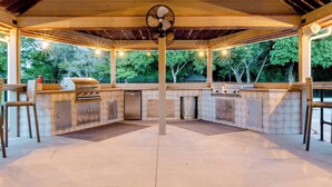 An outdoor kitchen with a grill, refrigerator, and sink, surrounded by a covered area with a fan and natural light. - Cozy Rustic Cabins for Self or Family Retreat (Whitewright)