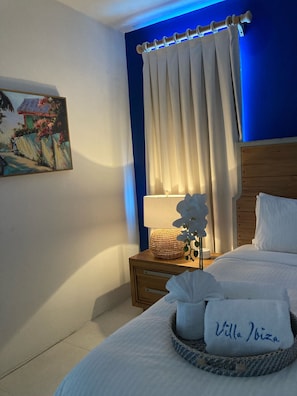 Quarto de hotel com cama, mesa de cabeceira, luminária, pintura na parede e janela com cortinas.