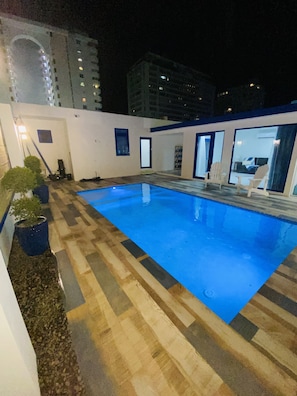 Piscina com deck de madeira, plantas em vasos e parede branca com janelas.