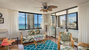 Ein geräumiges Wohnzimmer mit großem Fenster, das Blick auf das Meer und die Stadtkulisse bietet, mit Deckenventilator und Möbeln im tropischen Stil.