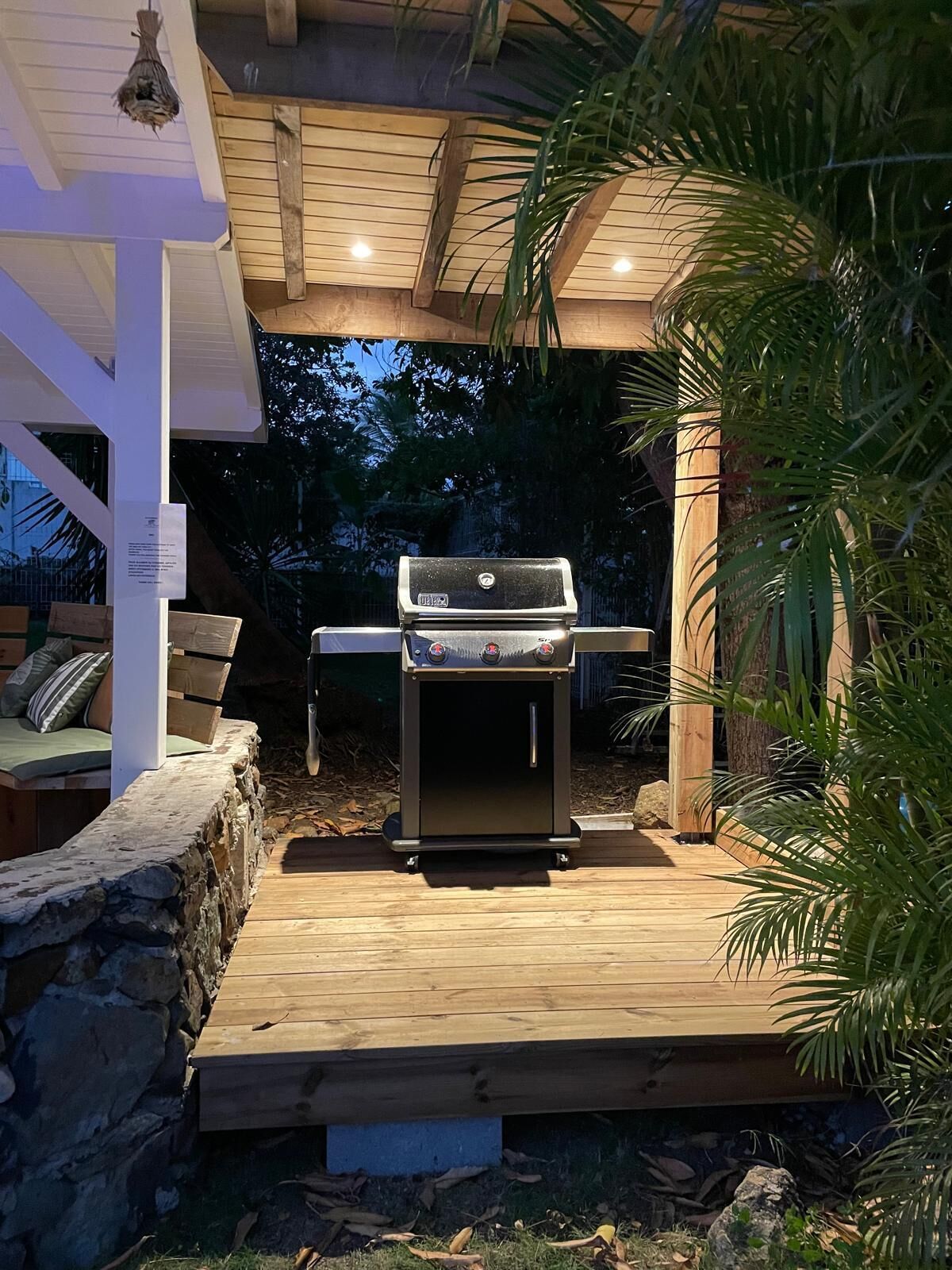 An outdoor grill area with a stone wall and wooden deck, under a covered structure with a sloped roof.