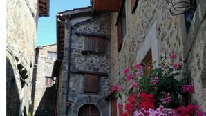 Ein schmaler Steinweg mit rustikalen Gebäuden, hölzernen Fensterläden und einem Blumenbeet mit rosa Blüten.