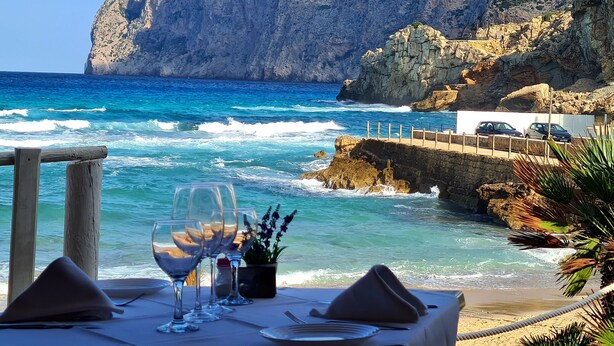 Une table dressée pour deux personnes, avec des verres à vin et une petite plante, donnant sur une côte rocheuse et la mer.