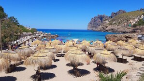 Une plage bordée de parasols en chaume, une côte rocheuse et des bâtiments construits sur une colline.