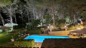 Vue nocturne d’un espace piscine avec un mur en pierre, des chaises longues, des parasols et des arbres.