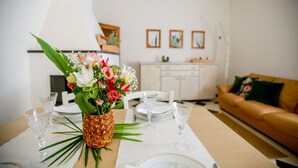 Et spisebord dækket med glas og en ananas som midtpunkt, et hvidt skab og en brun lædersofa i et rum med hvide vægge og billeder i rammer.