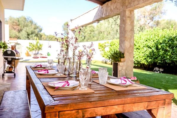 An outdoor wooden table set with plates, cutlery, and clear glasses, surrounded by potted plants and a barbecue grill.