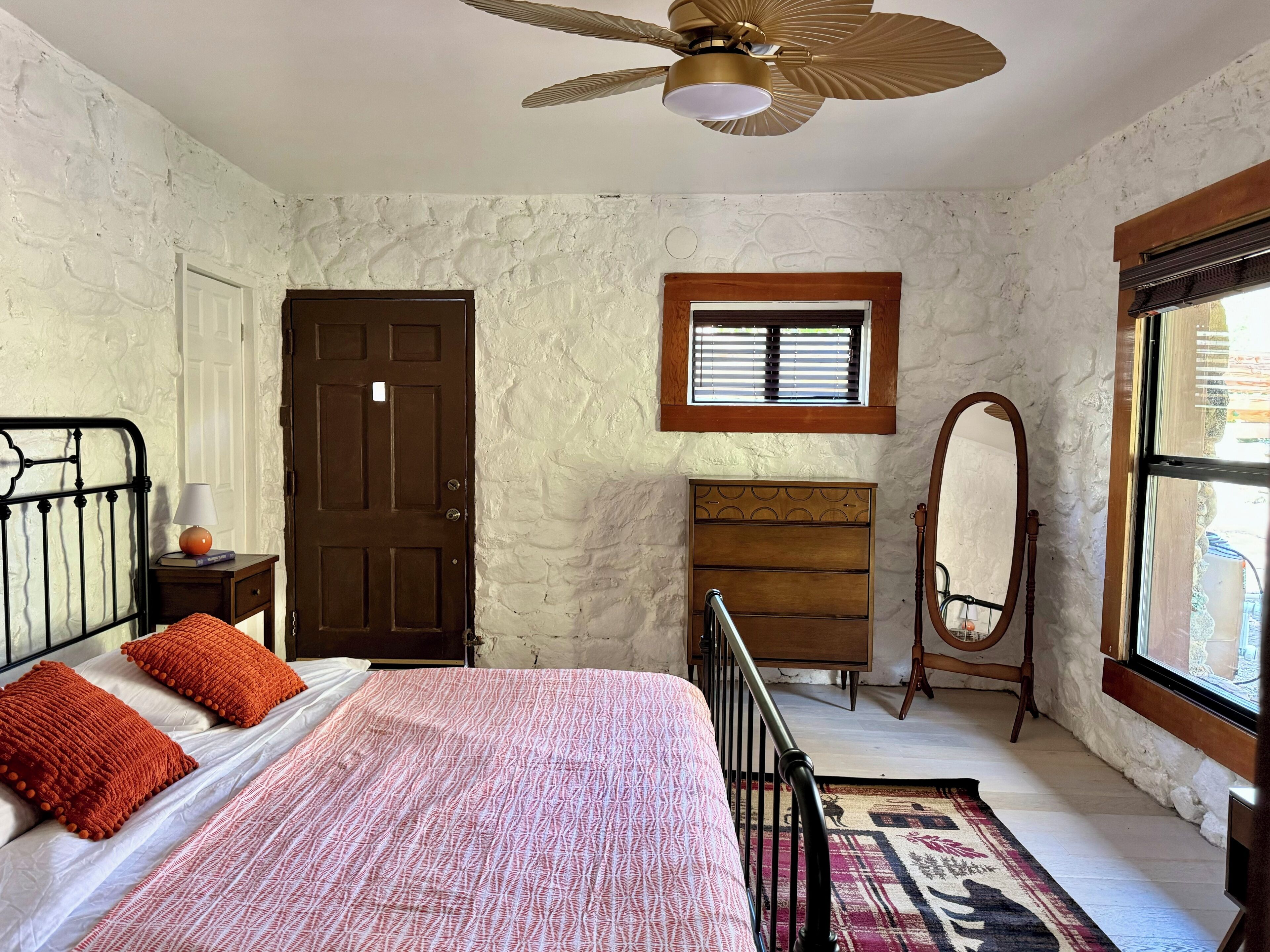 Une chambre à coucher comprenant un lit, une commode en bois, un miroir, un ventilateur de plafond et une fenêtre avec des stores.
