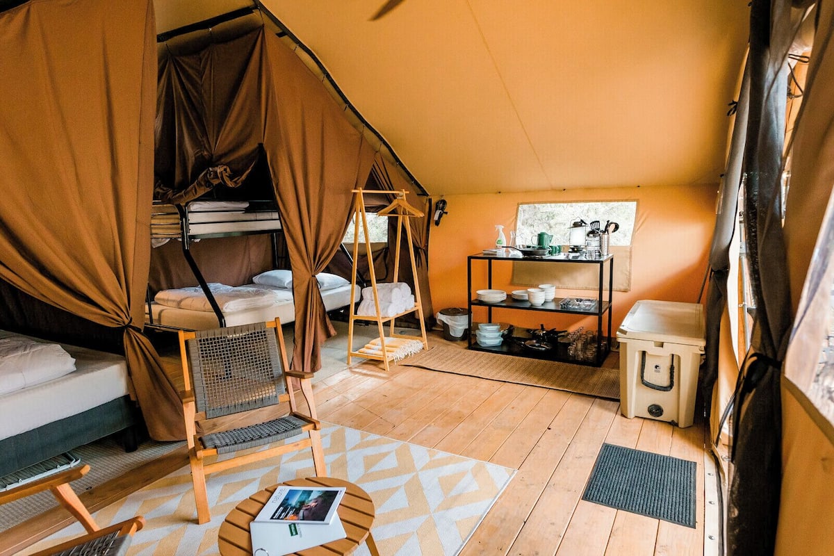 Alojamiento con carpa que incluye una cama, una silla colgante, un estante con vajilla y una mesita con una revista.