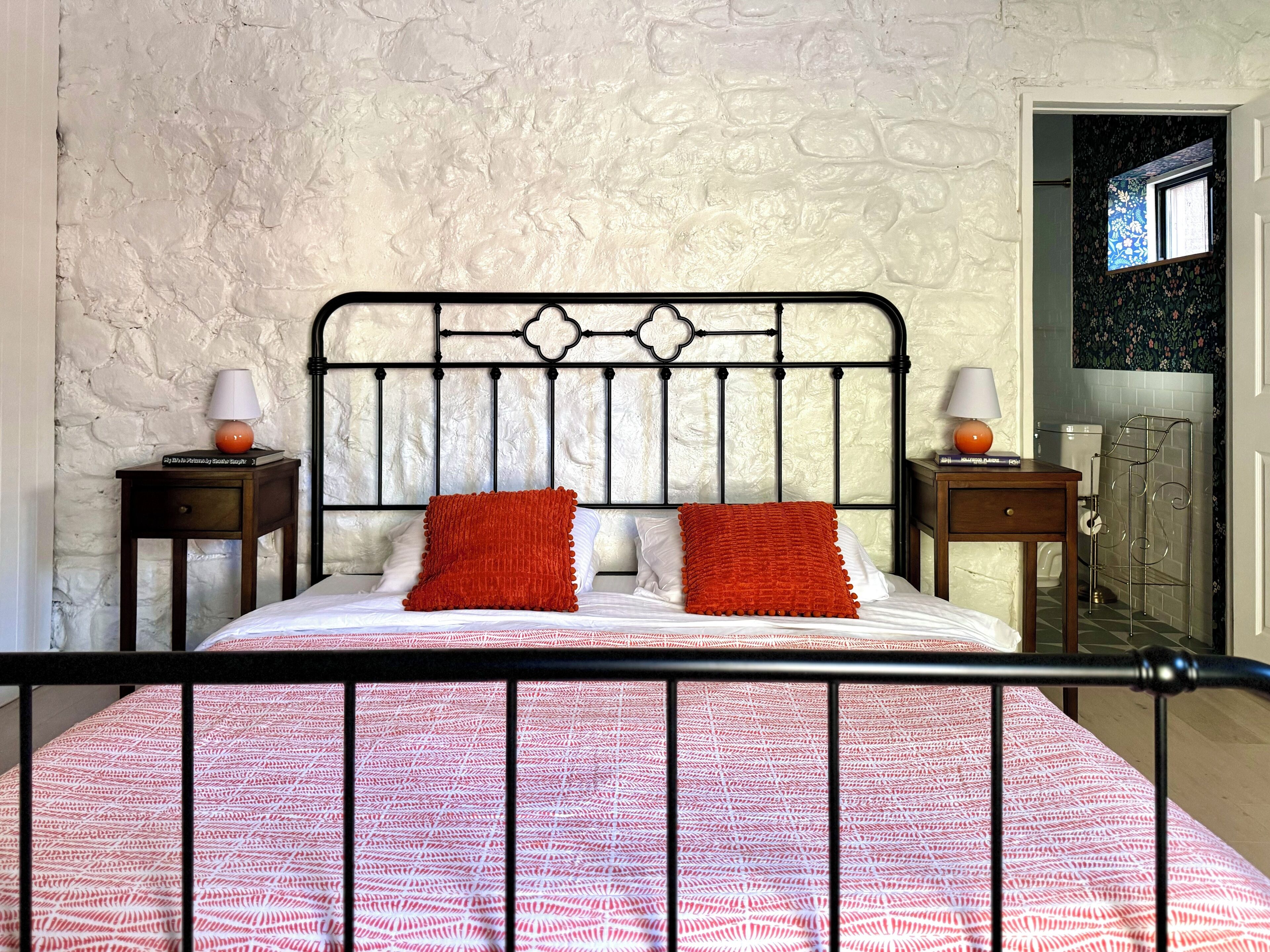 A bed with a patterned bedspread, two pillows, and a metal headboard. There are two bedside tables with lamps and a small book on one. The room has a textured wall and a window with a view of a bathroom.