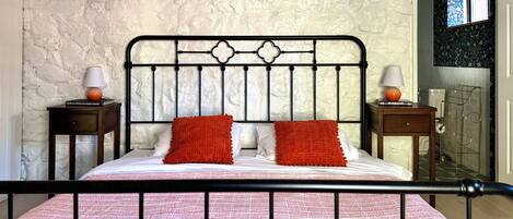 A bed with a patterned bedspread, two pillows, and a metal headboard. There are two bedside tables with lamps and a small book on one. The room has a textured wall and a window with a view of a bathroom.