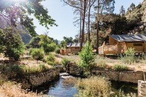 A rustic cabin with a stone bridge over a stream, surrounded by trees and a mountainous backdrop. - Huttopia Paradise Springs (Valyermo)